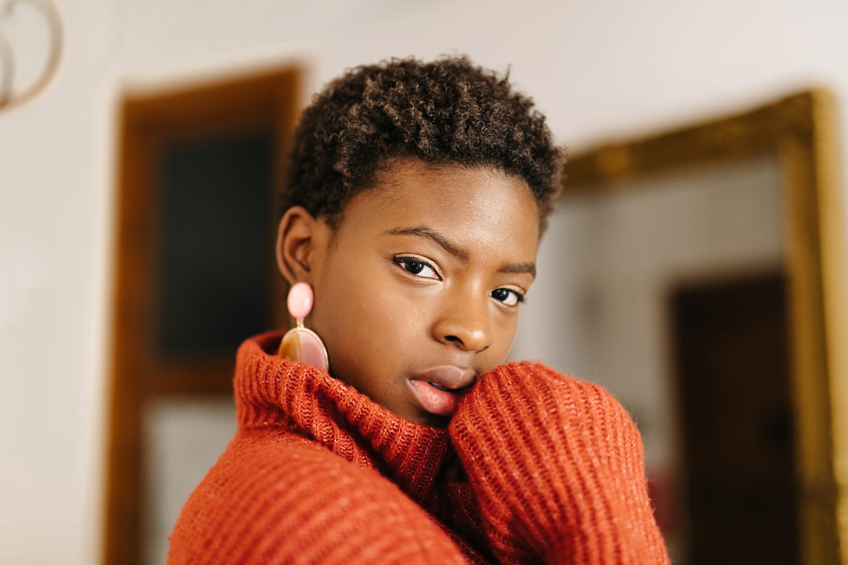 Lovely African American female looking at camera in orange wool sweater touching face while standing near open window in cozy apartment