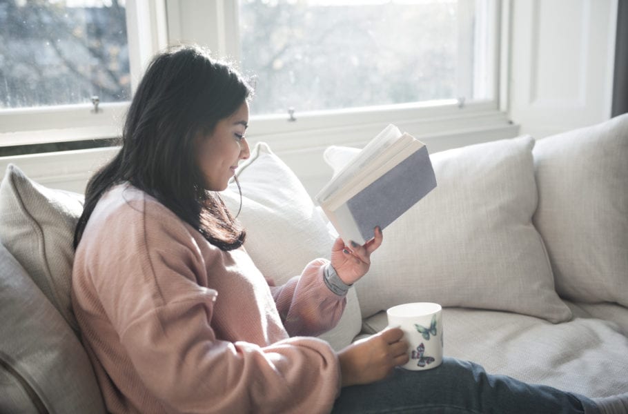 A British-Bangladeshi woman smiles as she reads a book and has a cup of tea while sitting on a white sofa next to a window in a flat in Edinburgh, Scotland, United Kingdom