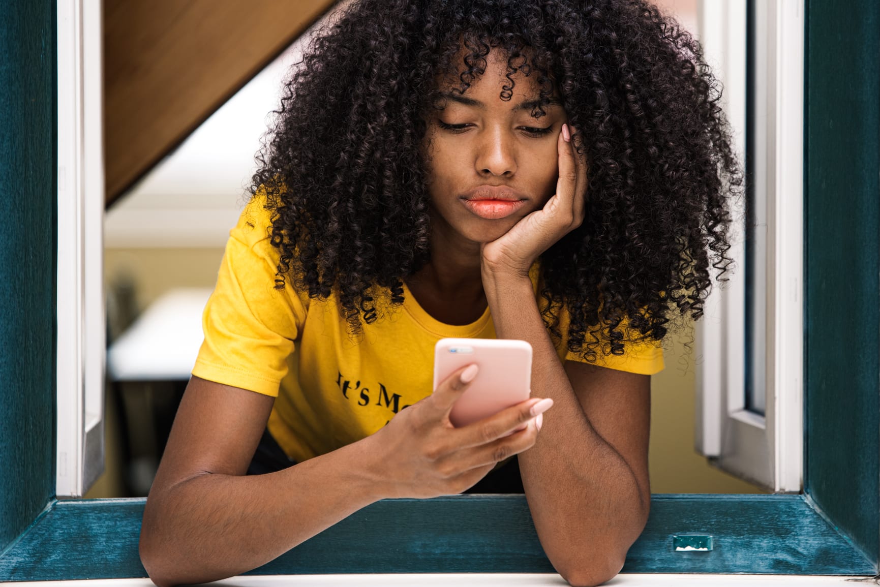 Pensive African American female with dark curly hair in bright yellow shirt looking out window and thinking while using smartphone in modern apartment