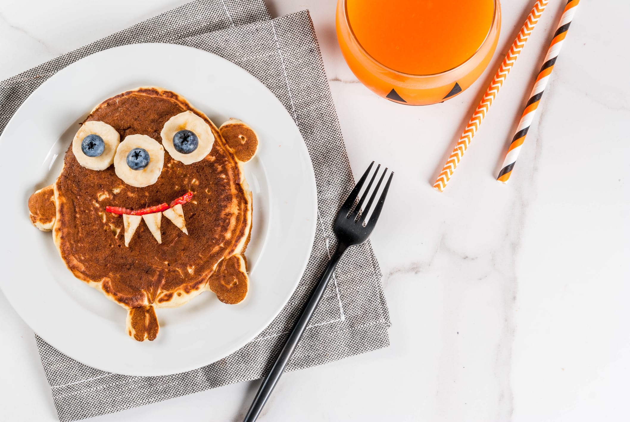 Funny food for Halloween. Kids breakfast pancake decorated like creepy monster, with banana, berries, with pumpkin smoothie juice, white table copy space top view