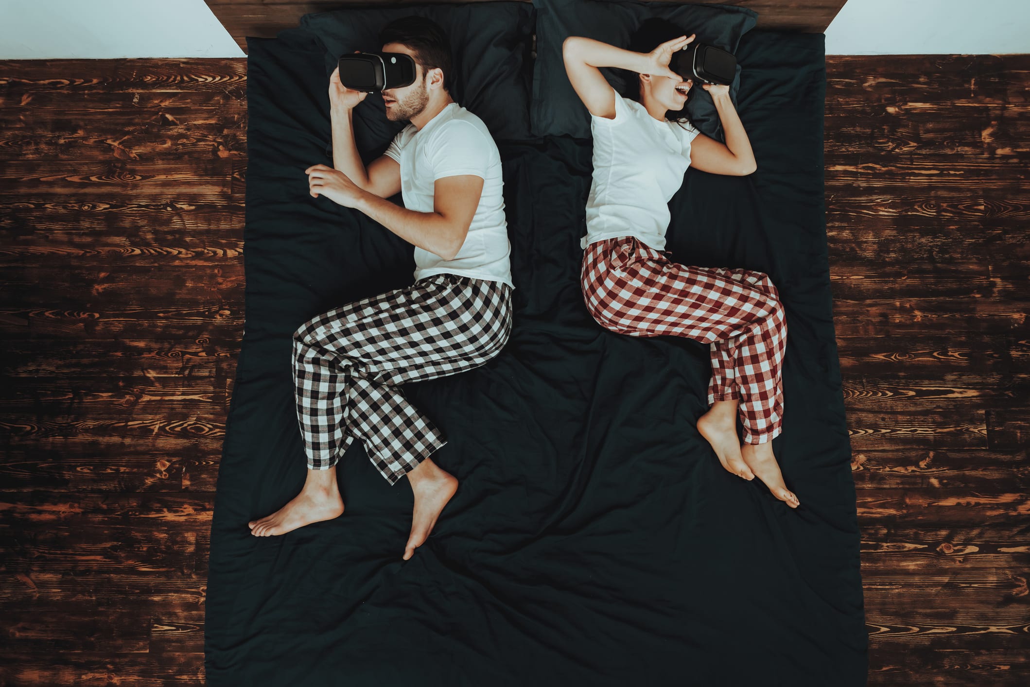Couple is Lying on Bed. Couple is Young Beautiful Woman and Man. People is Using Virtual Reality Glasses. People is Wearing Pajama Pants and T-Shirts. Persons in Home Interior. Top View.
