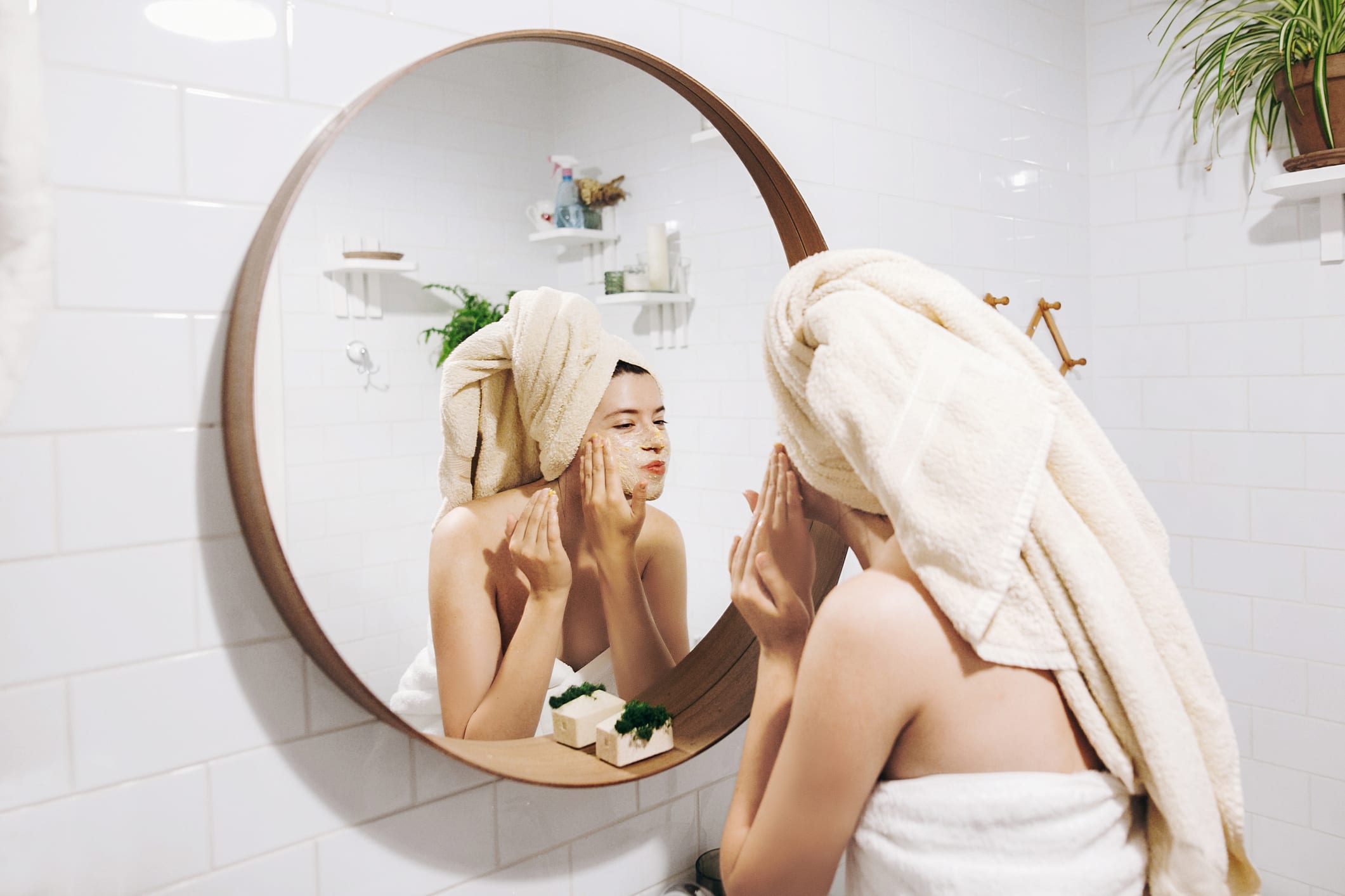 Young happy woman in towel making facial massage with organic face scrub and looking at mirror in stylish bathroom. Girl applying scrub cream, peeling and cleaning skin. Skin Care