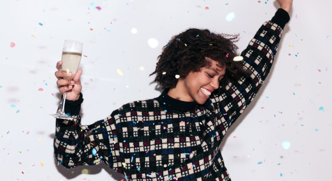 Happy mixed race woman with afro hair, wearing a dark plaid jumper, dancing and celebrating a party with confetti and a glass of champagne.