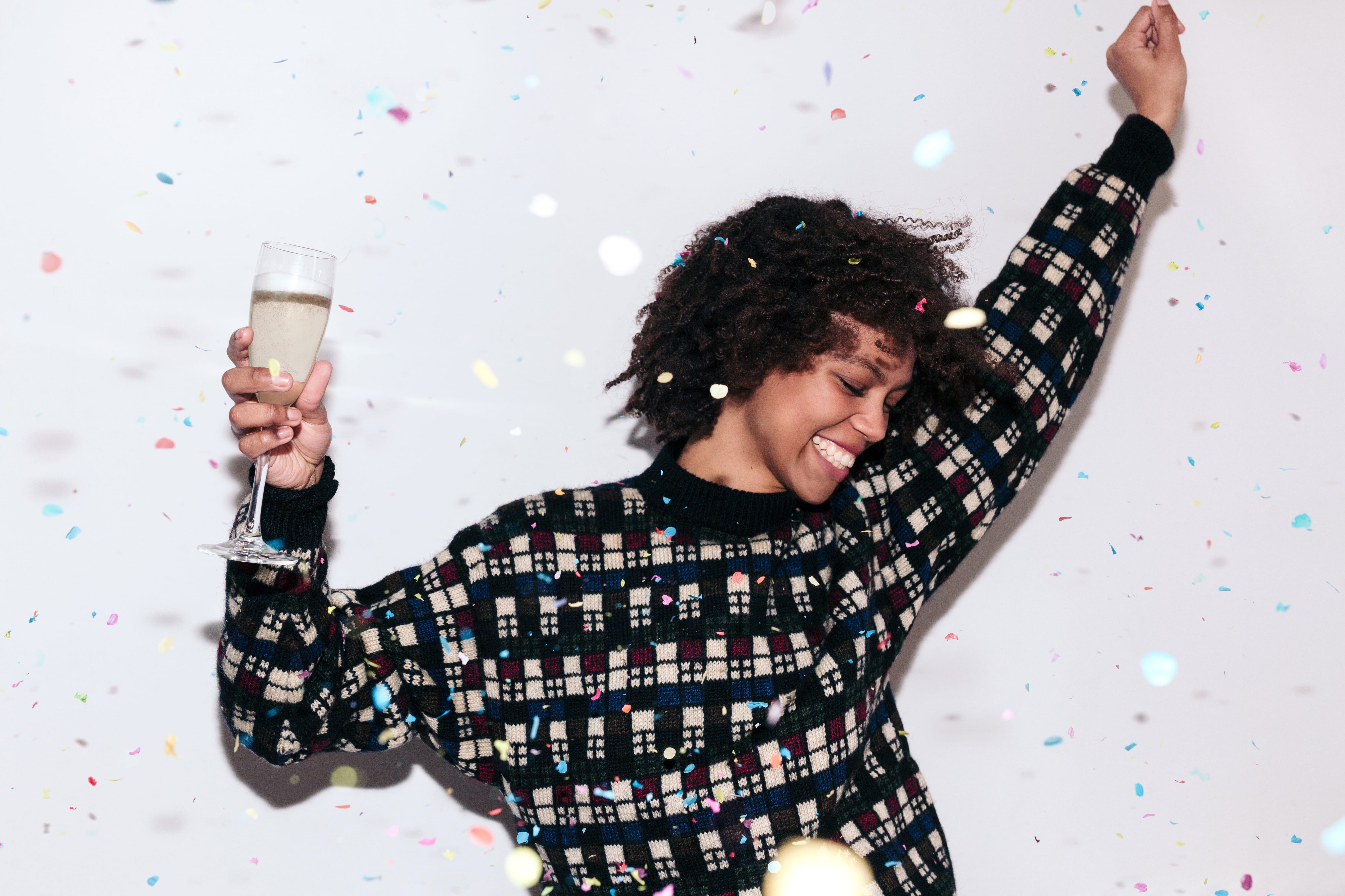 Happy mixed race woman with afro hair, wearing a dark plaid jumper, dancing and celebrating a party with confetti and a glass of champagne.