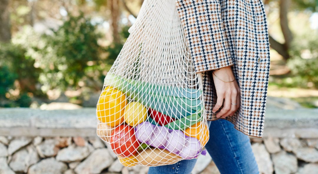 Woman carrying a reusable mesh shopping bag full of healthy fresh vegetables while walking along a sidewalk