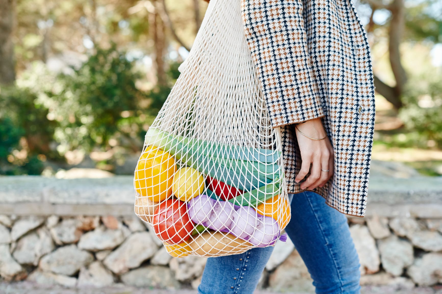 Woman carrying a reusable mesh shopping bag full of healthy fresh vegetables while walking along a sidewalk