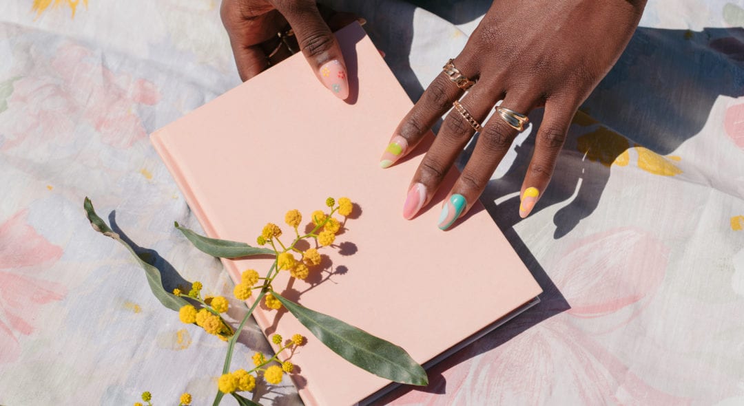 Black female hands with rings and colorful nails, holding a pastel pink notebook.