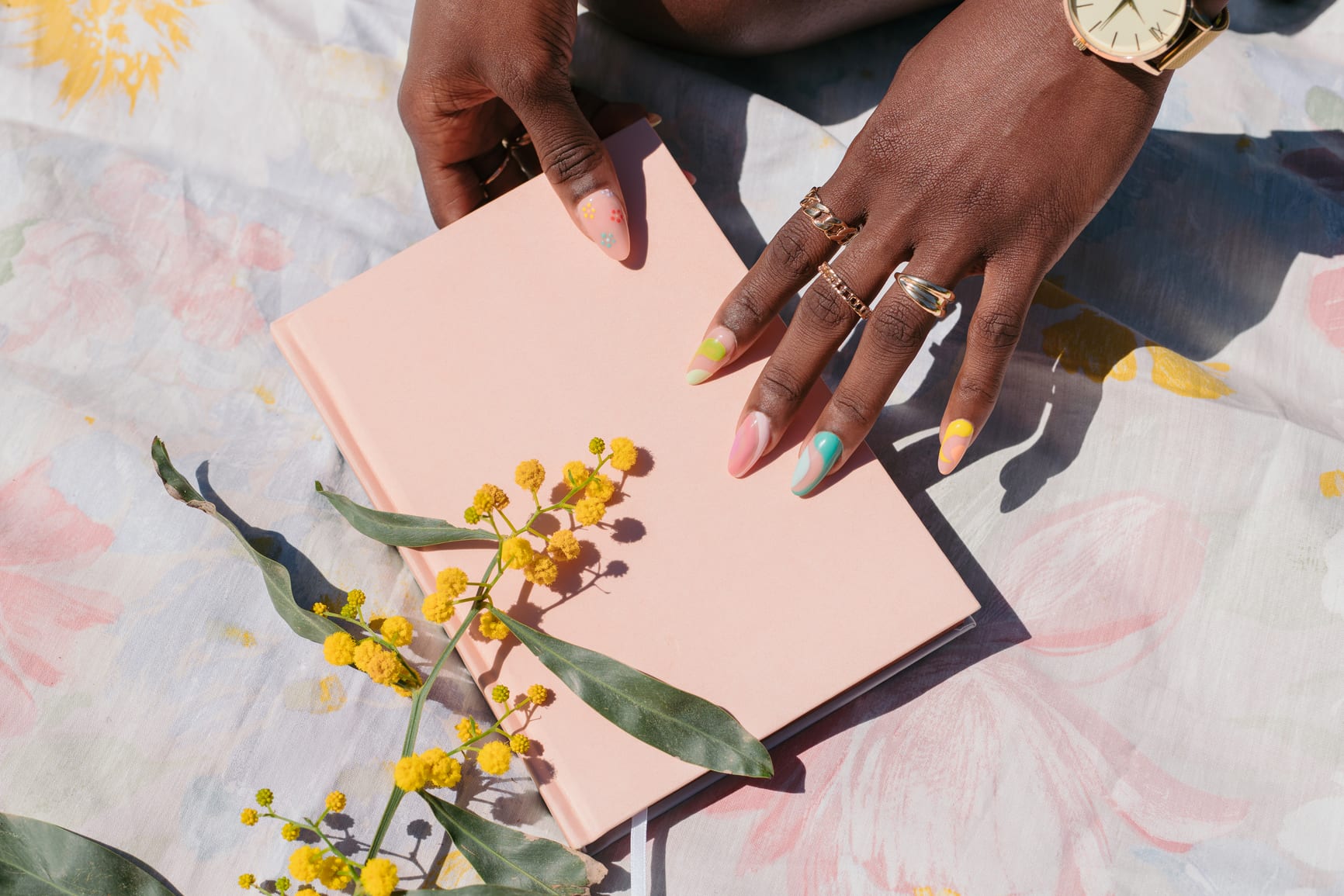 Black female hands with rings and colorful nails, holding a pastel pink notebook.