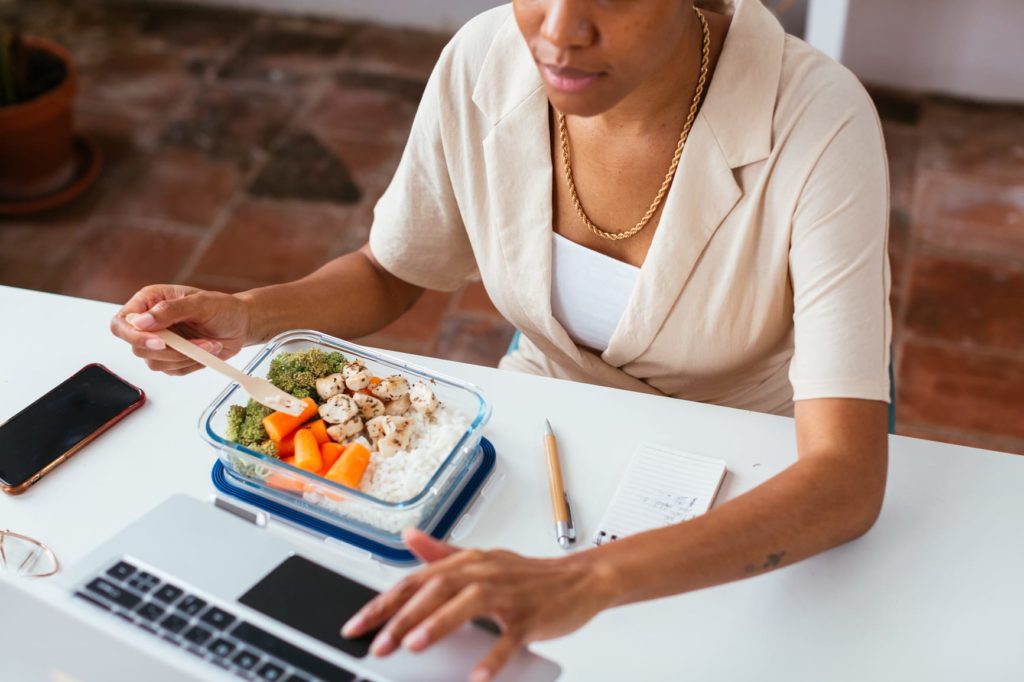 From above unrecognizable African American manager eating healthy food and browsing laptop while sitting at table during lunch in office