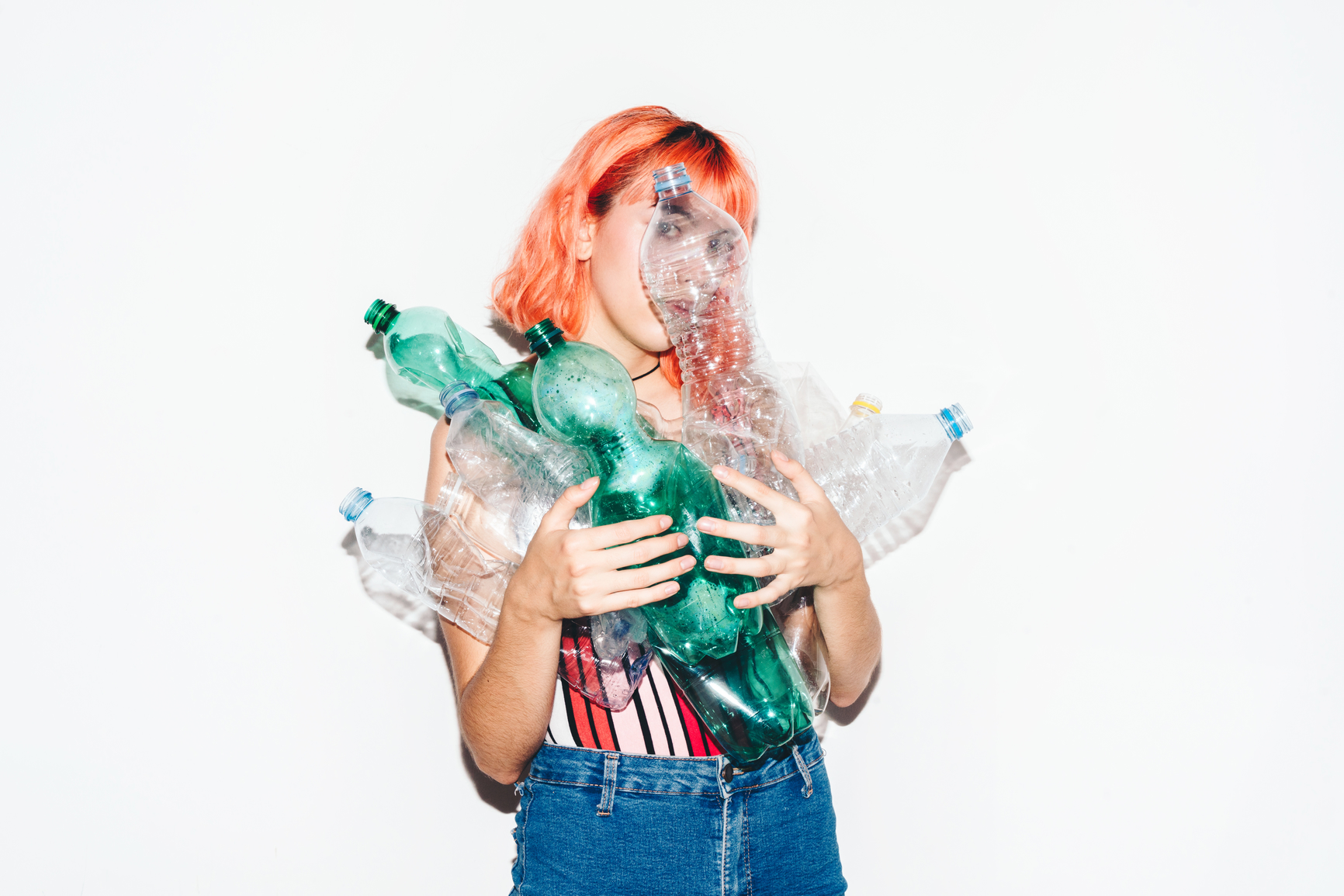 Young woman with red hair holding a bunch of empty single-use plastic bottles that cover her face. Recycling concept. White copy space in the background.