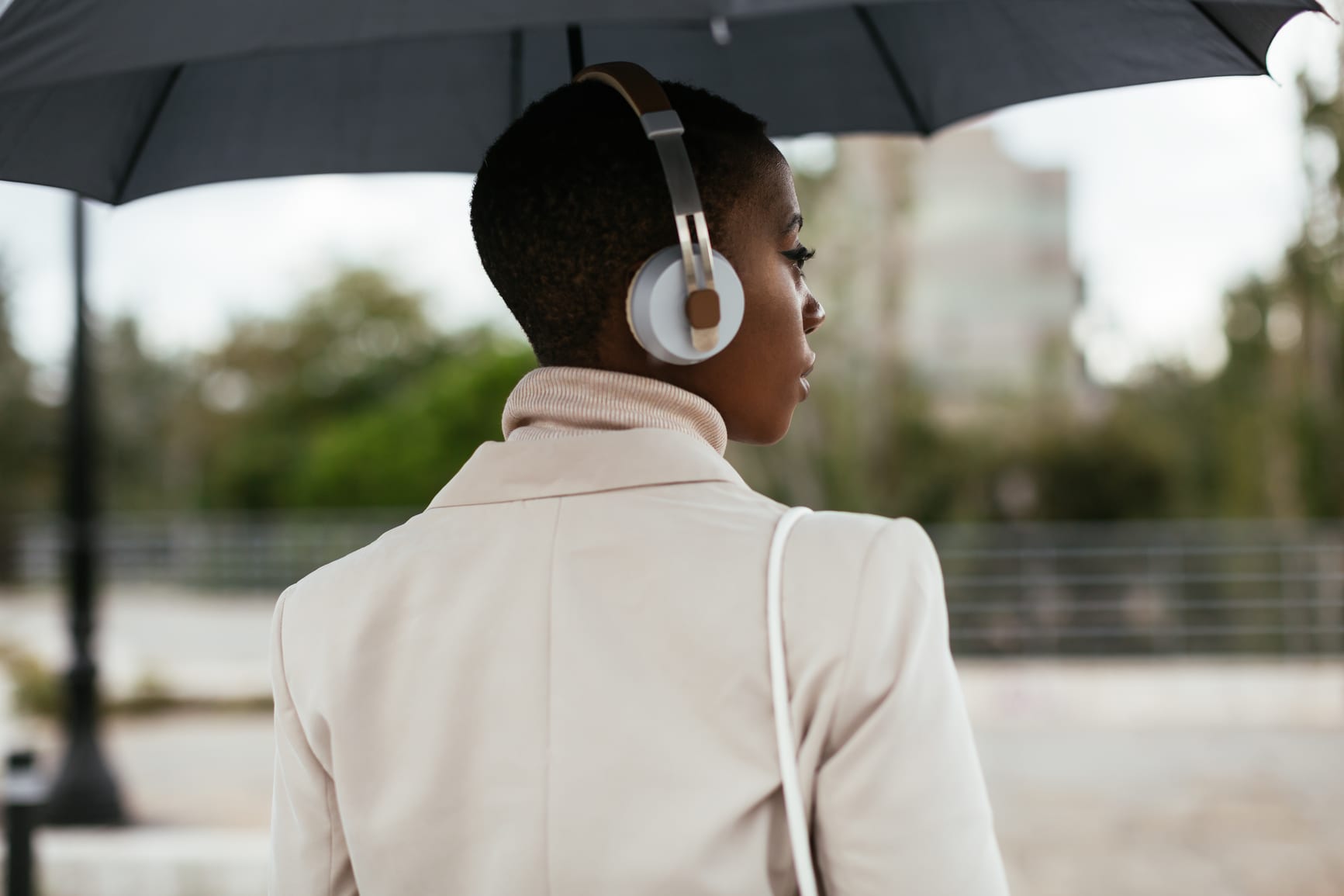 Back view of stylish African American woman in headphones listening to music and looking away while standing under umbrella on blurred background of city on rainy day