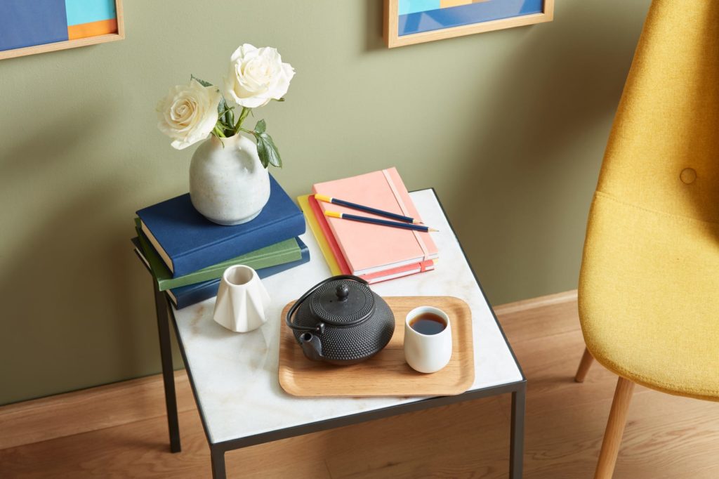 Top view of square marble table with colorful notebooks, pencils, books and flowers, and tray of fresh tea and a teapot beside modern yellow chair in daylight.