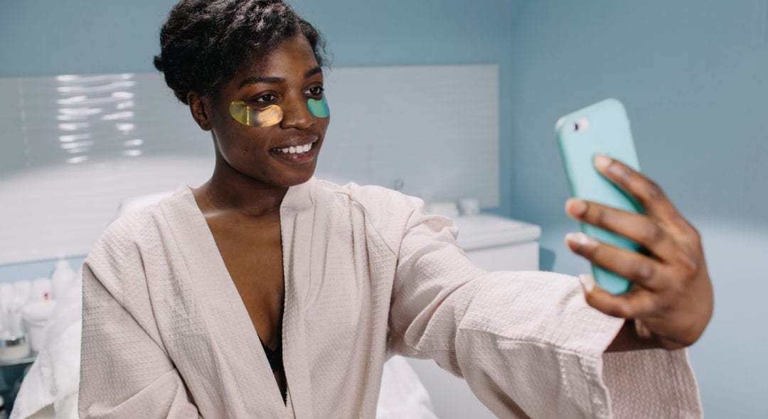 Cheerful black female with under eye patches smiling and taking selfie during skincare procedure in salon