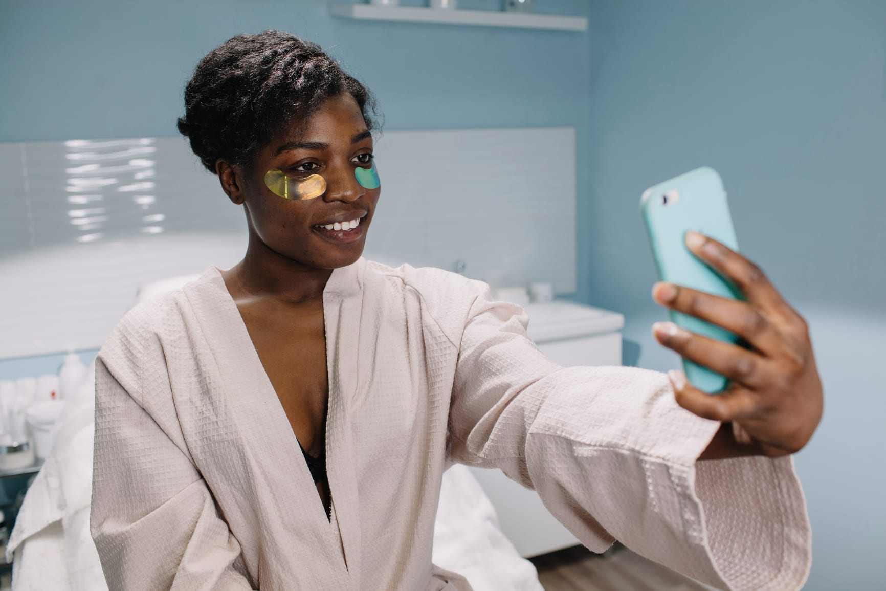 Cheerful black female with under eye patches smiling and taking selfie during skincare procedure in salon