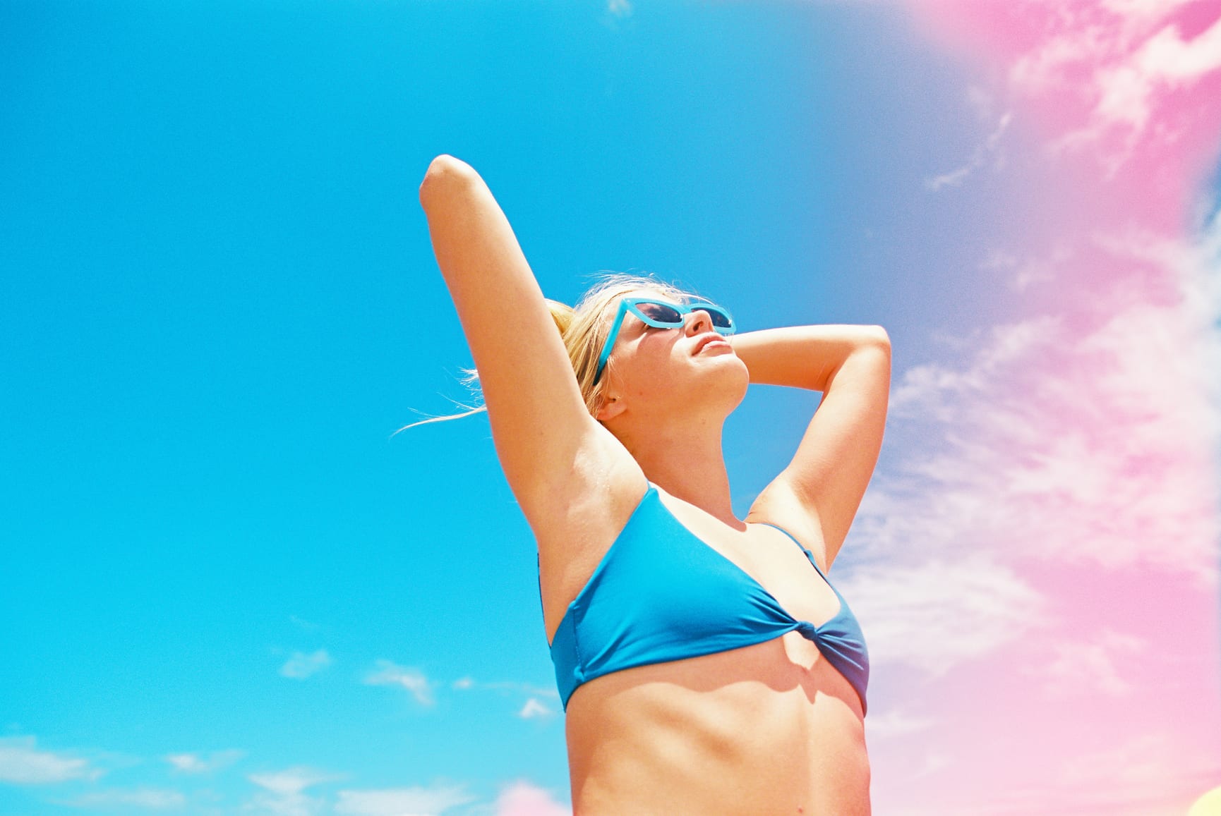 Blonde girl in blue bikini at beach in full sun in summer with a sunglasses and rainbow towel