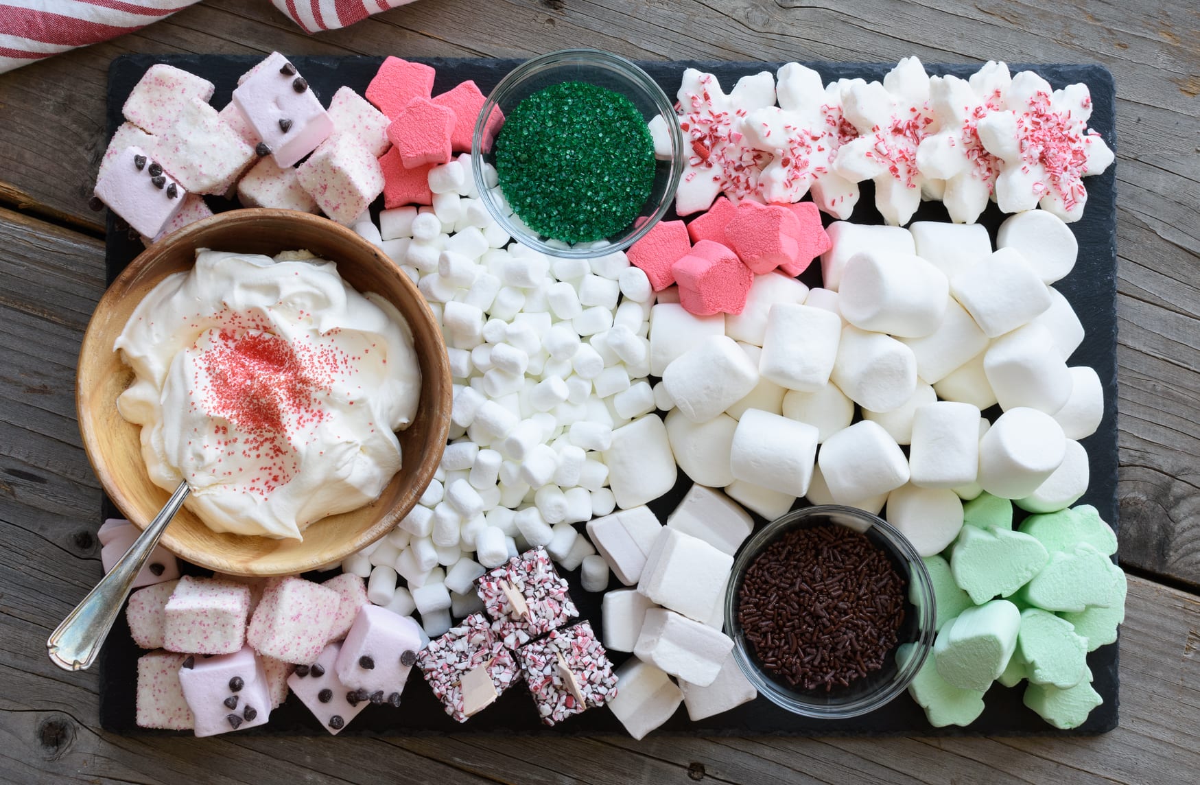 overhead shot of a variation on a charcuterie board, but with hot chocolate fixings instead of meats and cheeses. The board includes a variety of marshmallows, chocolate sprinkles, green sprinkles, chocolate and peppermint coated wooden spoons, and whipped cream