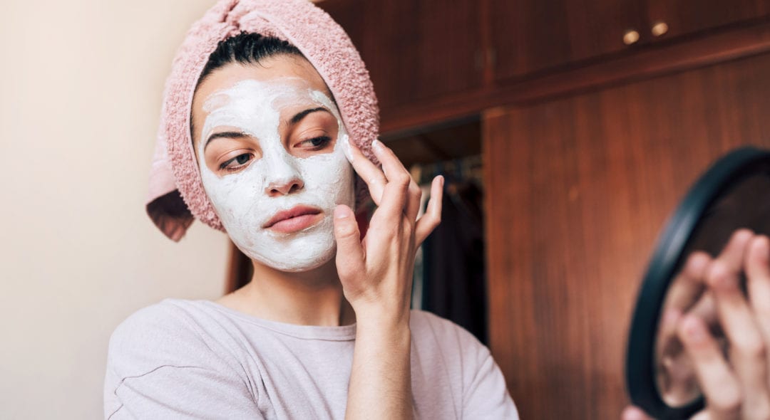 Young woman with a towel wrapped around her hair, applying a cleansing face mask at home.