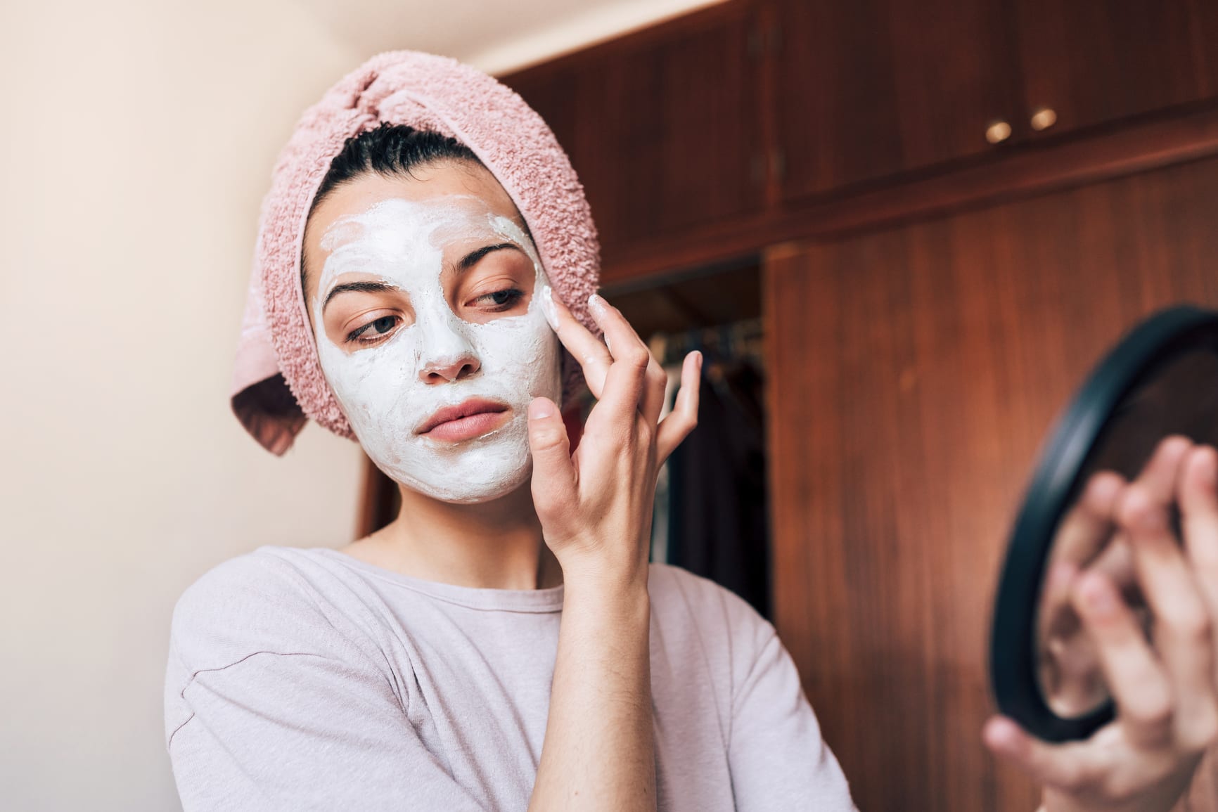 Young woman with a towel wrapped around her hair, applying a cleansing face mask at home.