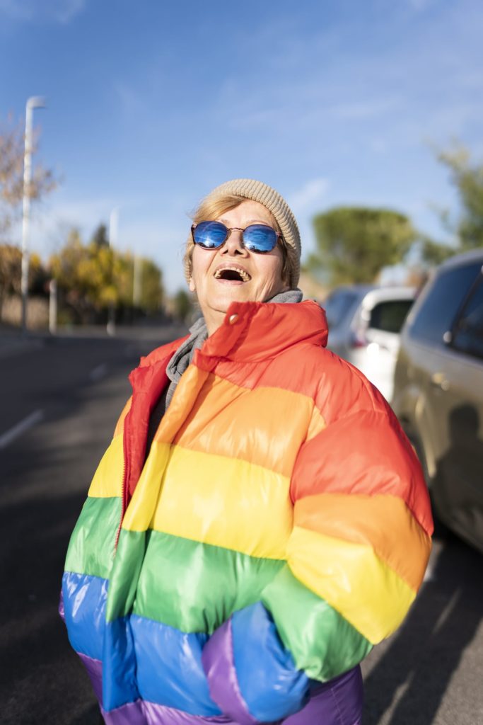 Grandmother in rainbow coat smiling in the middle of the street. A winter day. pride lgtb.