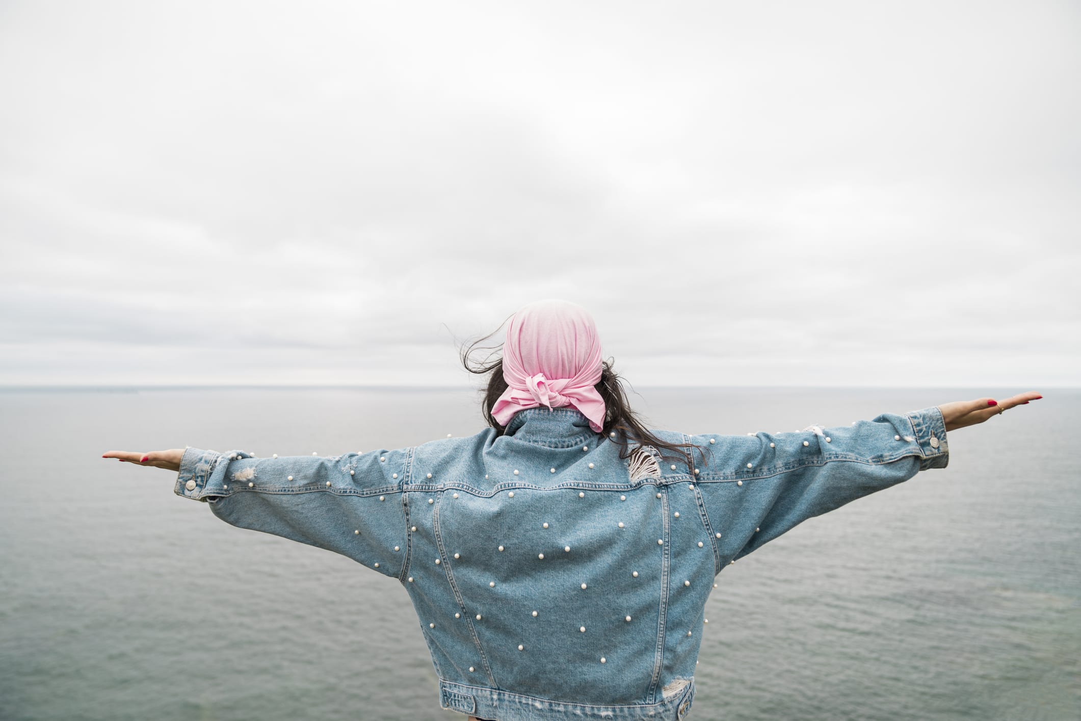 Young female cancer survivor with arms outstretched looking at sea against cloudy sky