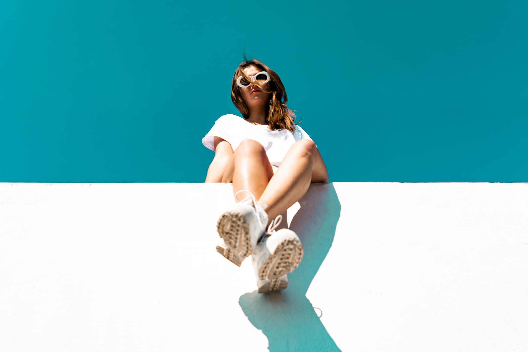 Young woman wearing large white sunglasses sitting on modern concrete wall. Simplicity Modern Urban Style. Shot from below against the sky. Urban Lifestyle Youth Portrait.