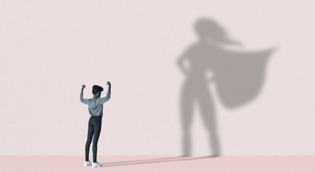 Young woman flexing muscles in front of large superhero shadow on pink background