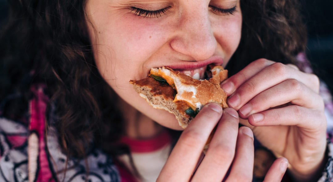 Close up of a Gen Z teenager eating a hamburger in her car, biting it aggressively.