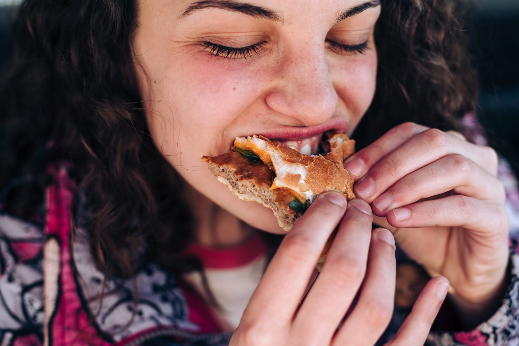 Close up of a Gen Z teenager eating a hamburger in her car, biting it aggressively.