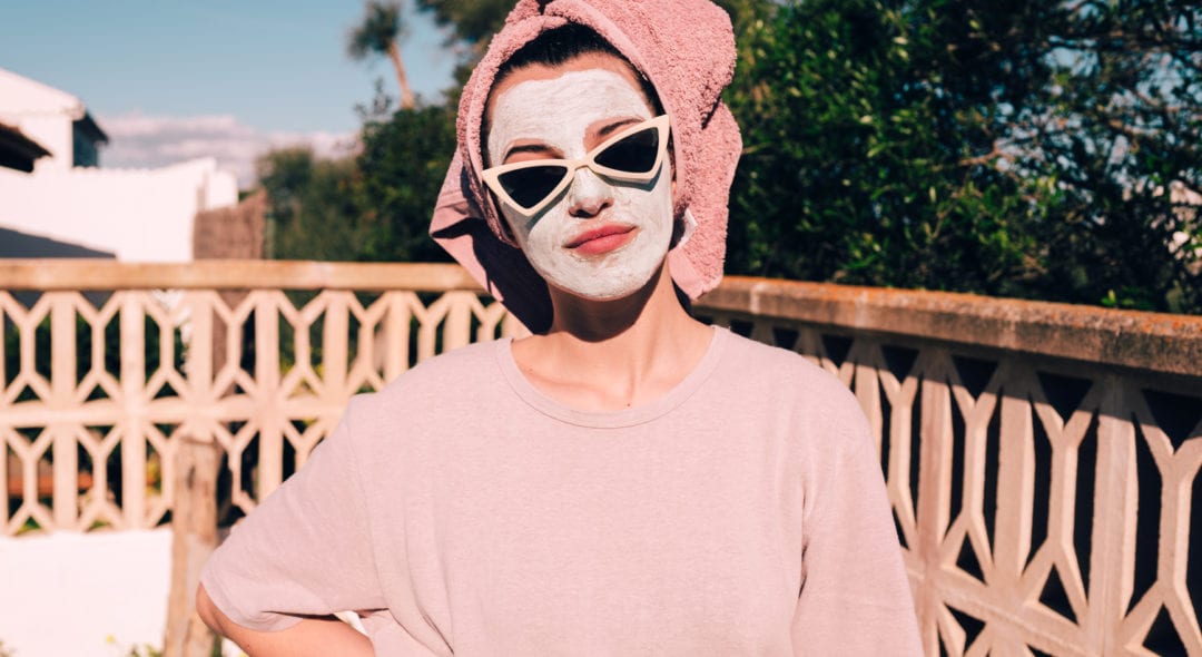 Young woman with white face mask and sunglasses, wearing a towel on her hair, looking at camera.