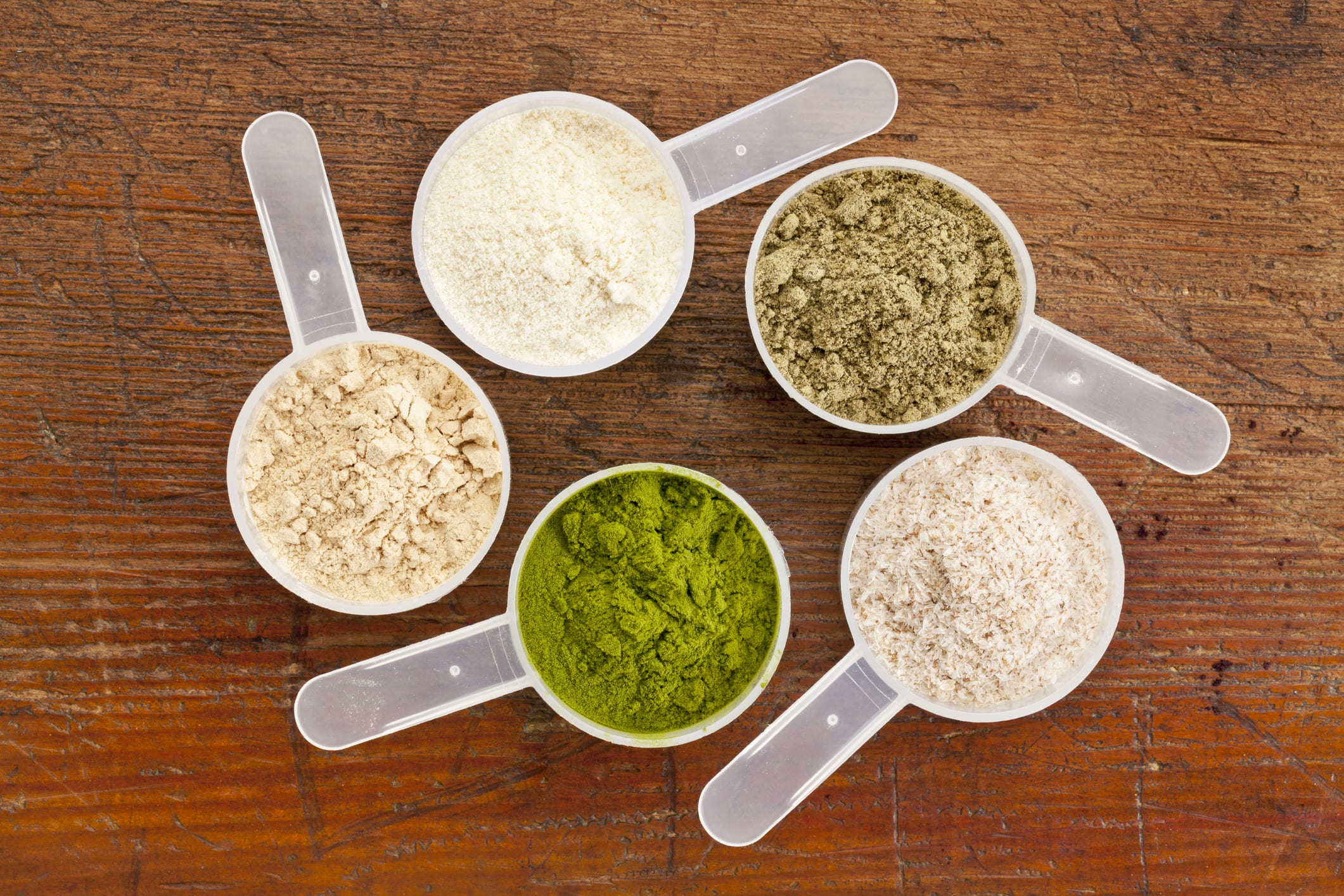 five plastic measuring cups of different superfood supplement powders (form bottom clockwise: wheatgrass, maca root, whey protein, hemp seed protein, psyllium husk) on grunge wood background