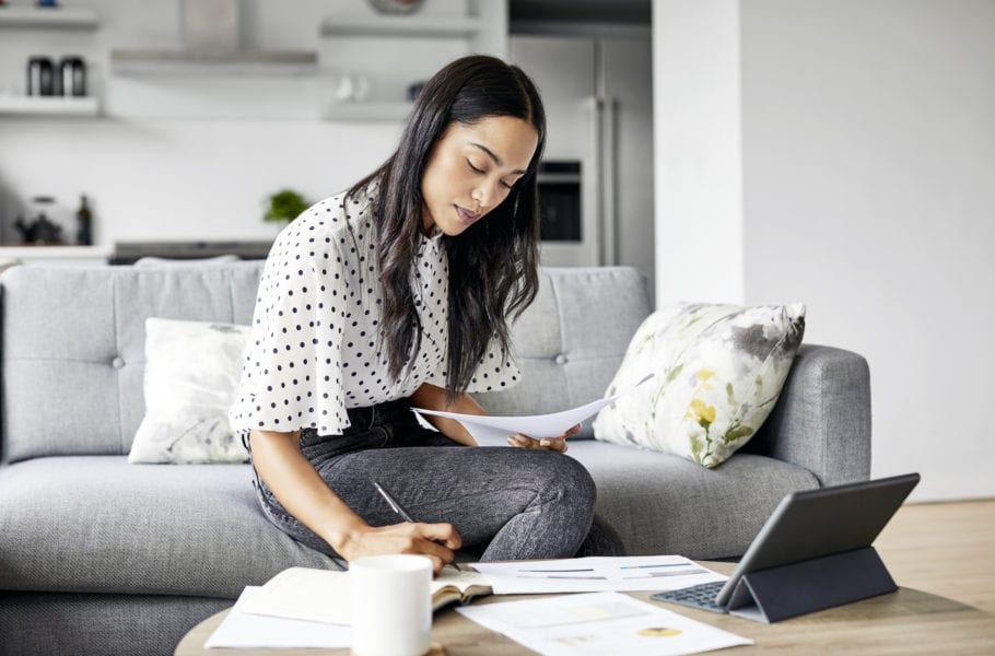 Young woman analyzing bills while writing in diary. Beautiful female is using digital tablet at table. She is sitting on sofa at home.