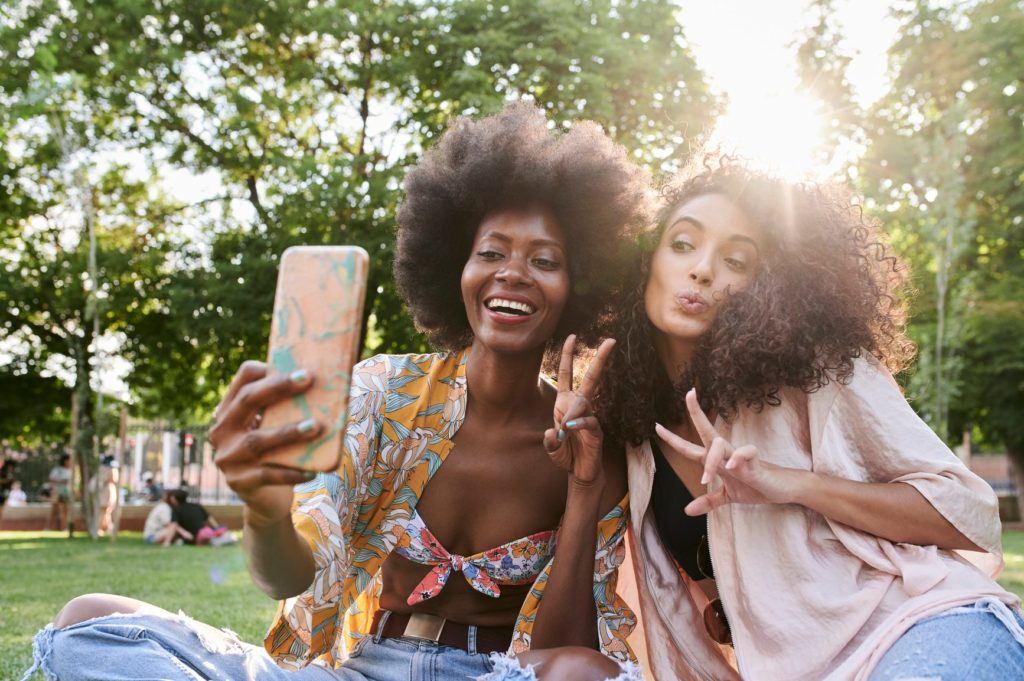 Casual millennial women best friends taking selfie with smartphone at park during sunset. Backlit.