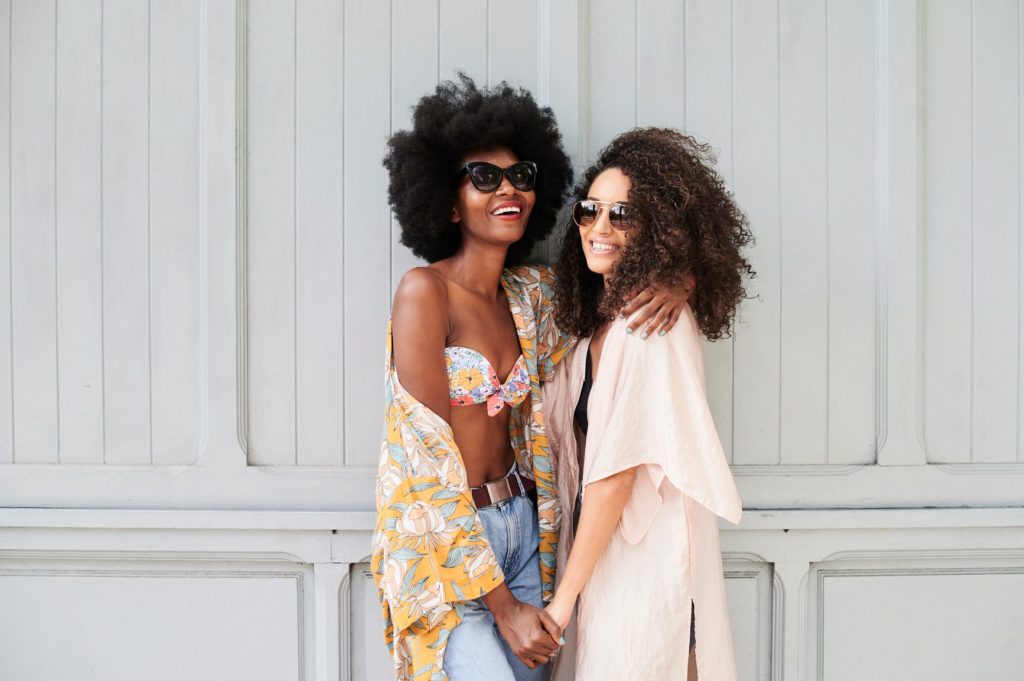 Millennial girls best friends portrait looking cheerful at camera embracing each other against wooden wall.
