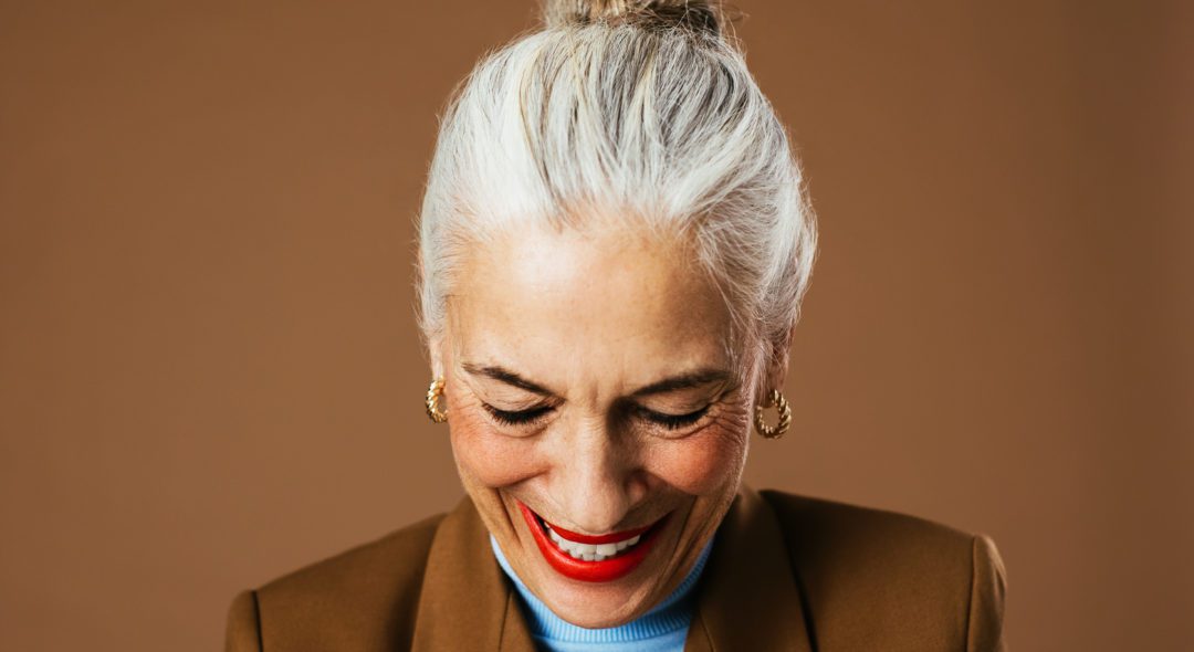 Happy elderly female in trendy jacket and turtleneck looking down and laughing against brown background