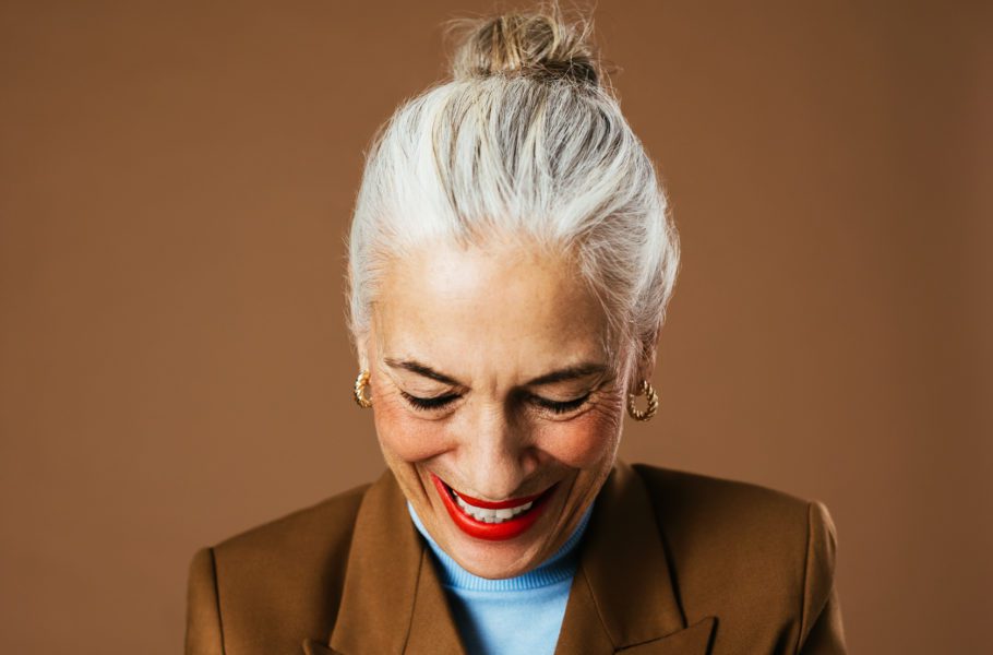 Happy elderly female in trendy jacket and turtleneck looking down and laughing against brown background