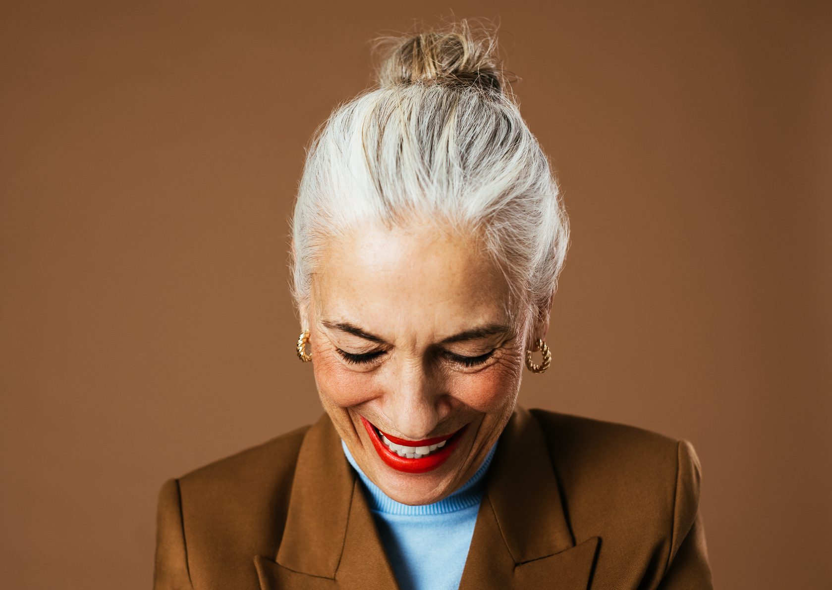 Happy elderly female in trendy jacket and turtleneck looking down and laughing against brown background