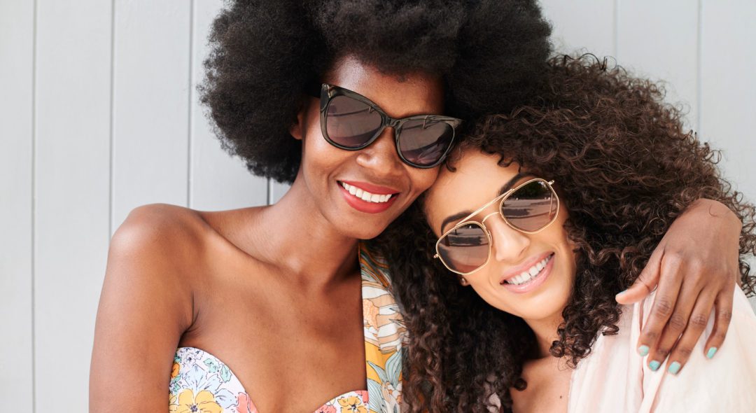 Multiethnic best friends portrait looking at camera wearing sunglasses in summer against grey background. Curly hair.