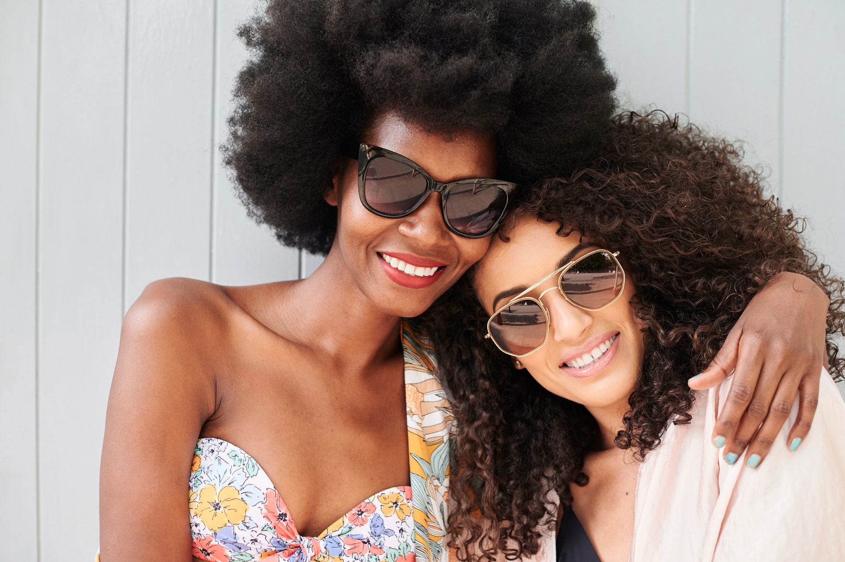 Multiethnic best friends portrait looking at camera wearing sunglasses in summer against grey background. Curly hair.