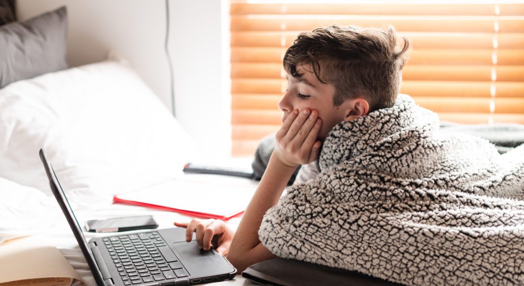 A young boy doing his school work in bed while school is on a break.