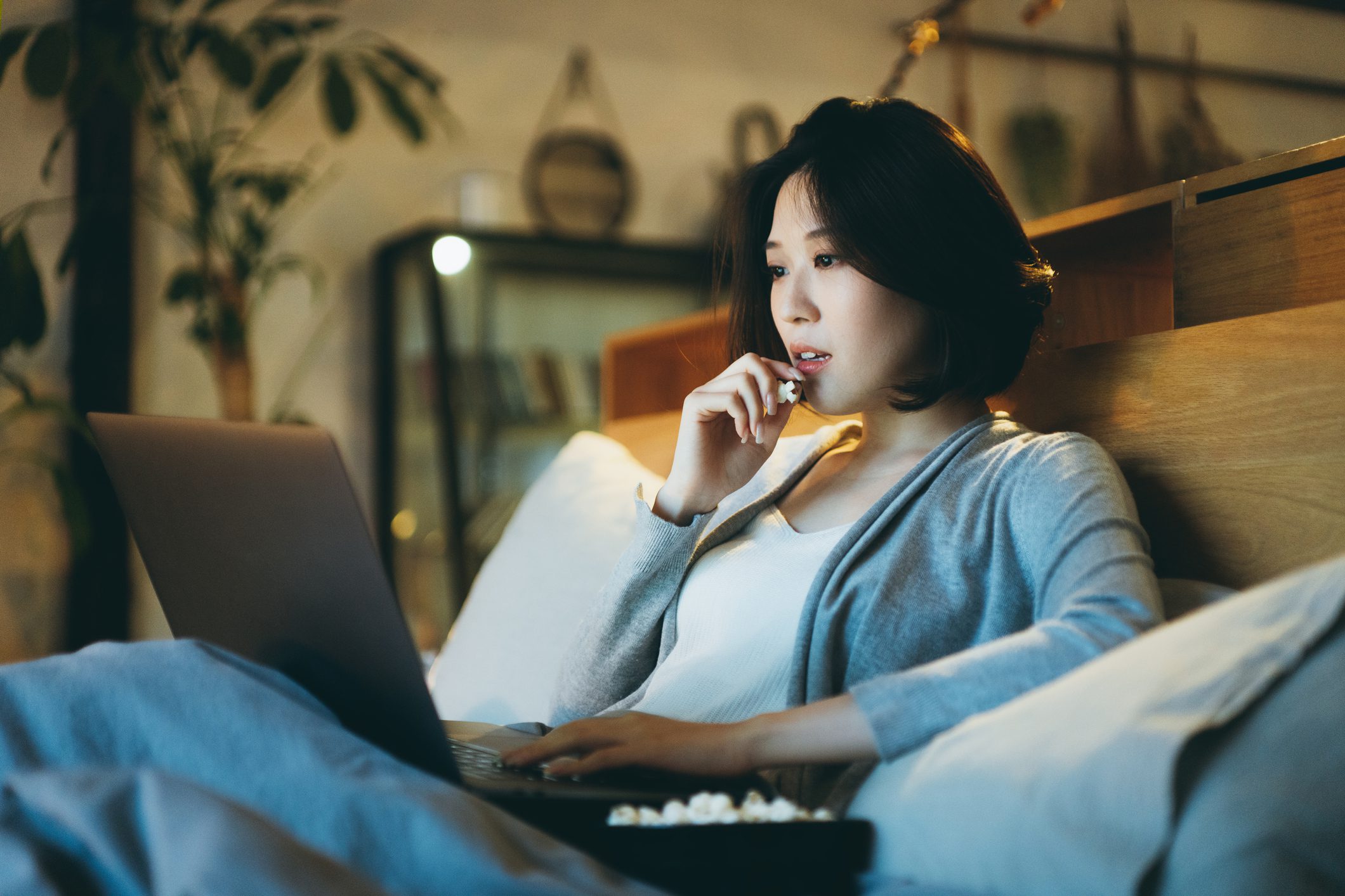 Young Asian woman lying on the bed enjoying the weekend, watching movie on laptop and eating popcorn at home in the evening