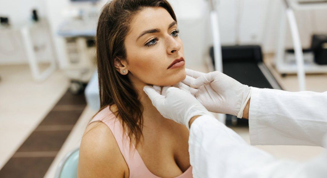Close-up of young woman visiting general practitioner due to soreness and pain in her throat. The doctor is touching her neck to see if there are any swollen glands