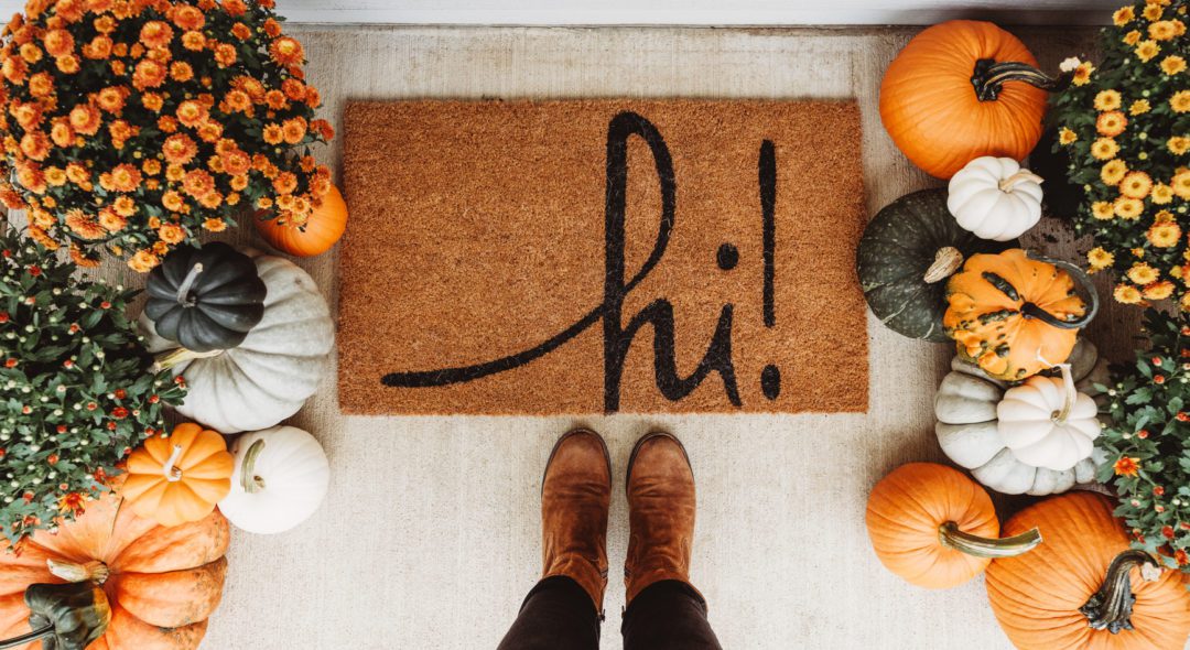 An entryway to a home with a Hi floor mat and pumpkins and mums decorated for fall season.