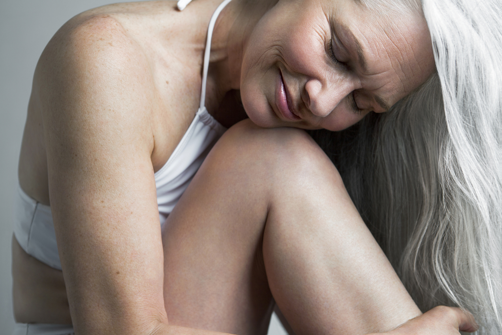 Mature woman in her late fifties in a white two piece swimsuit with long, white, silver grey hair hugging her knee with her eyes closed and a big smile, cropped.