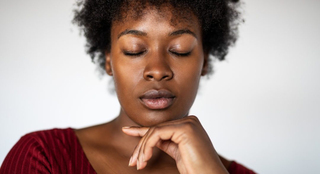 Close-up portrait of an african young woman with eyes closed against white background. Female standing with hand on chin.