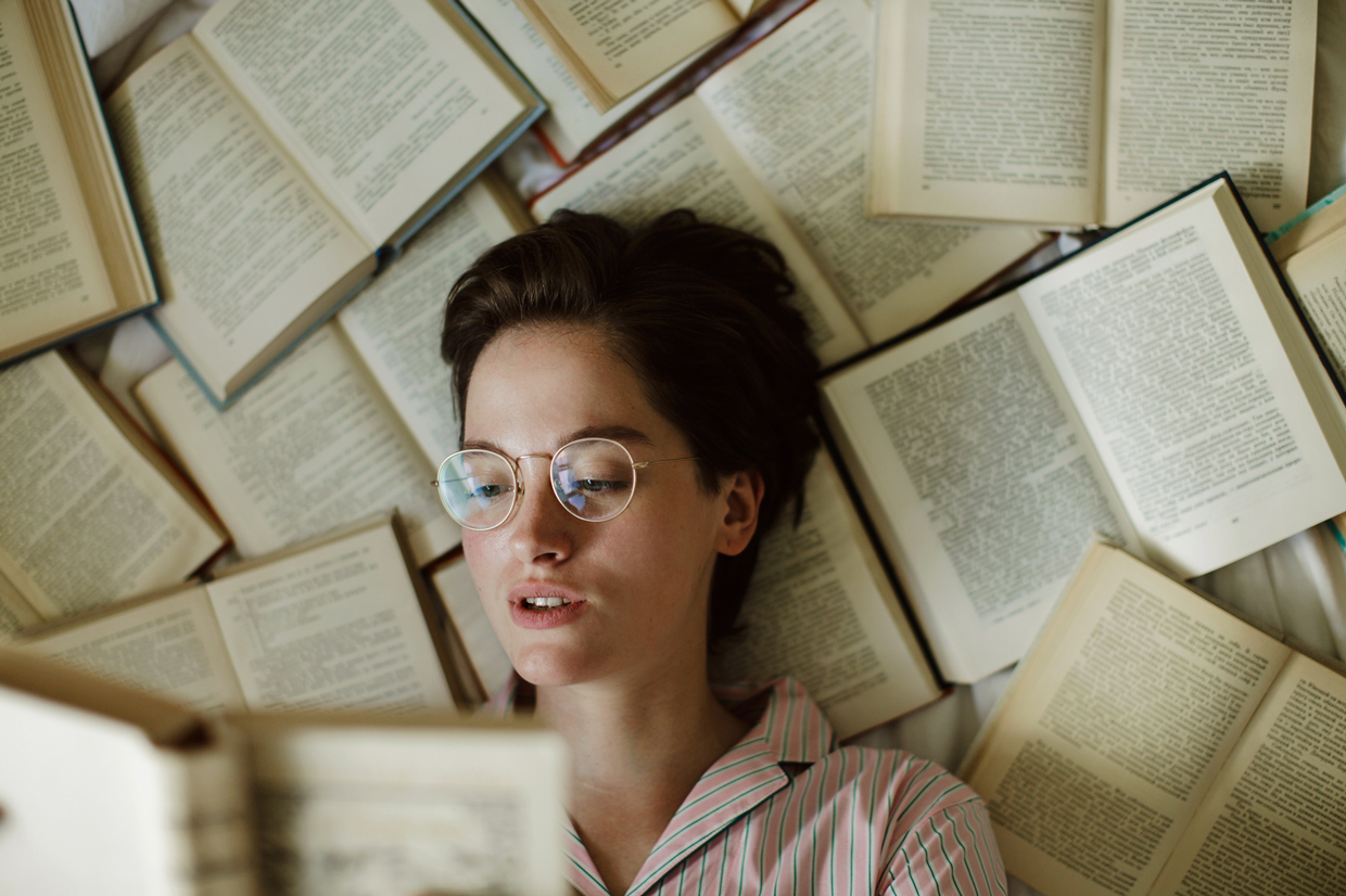 young woman with glasses laying on books reading a book