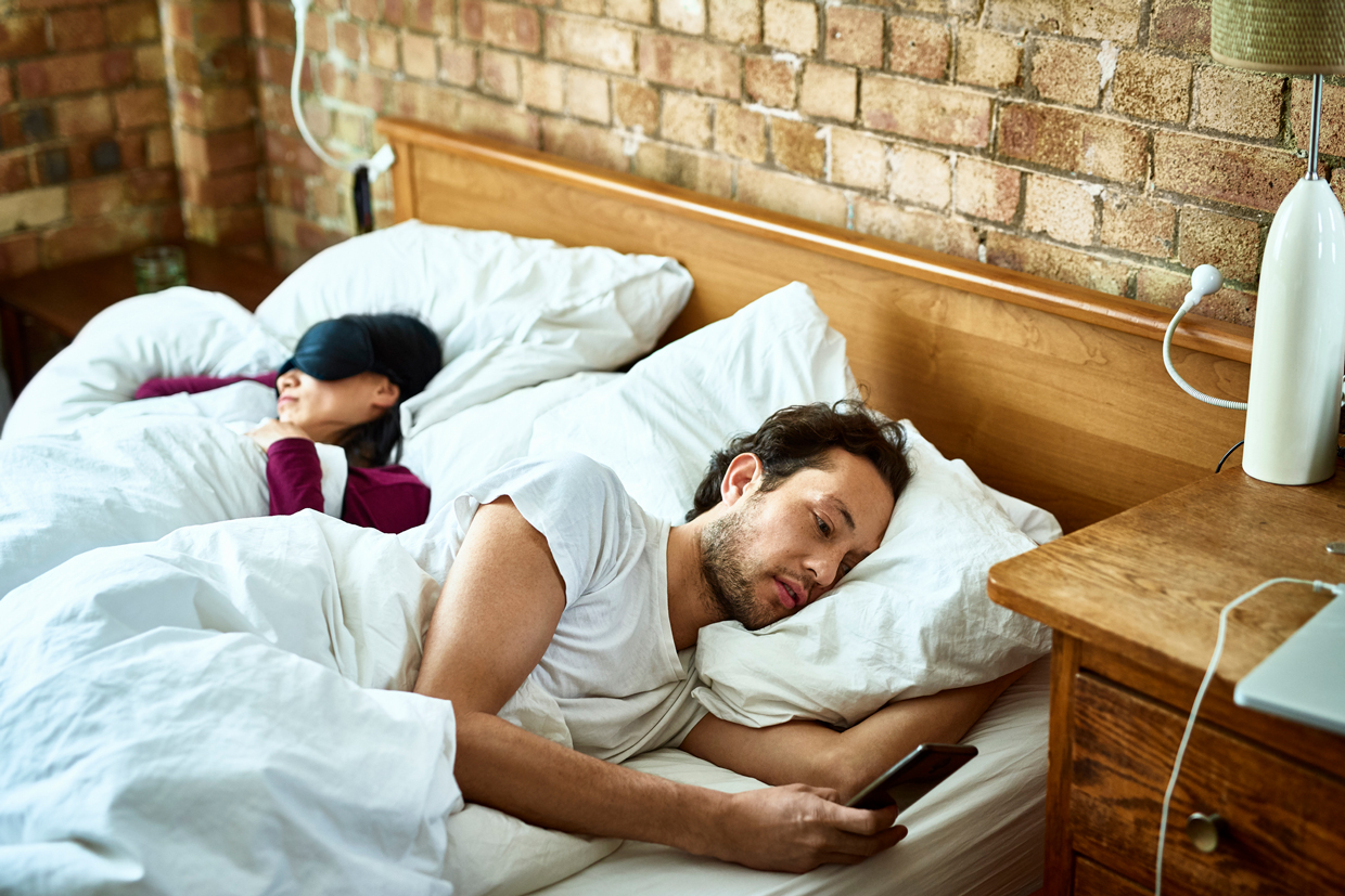 man in bed looking at phone with sleeping woman