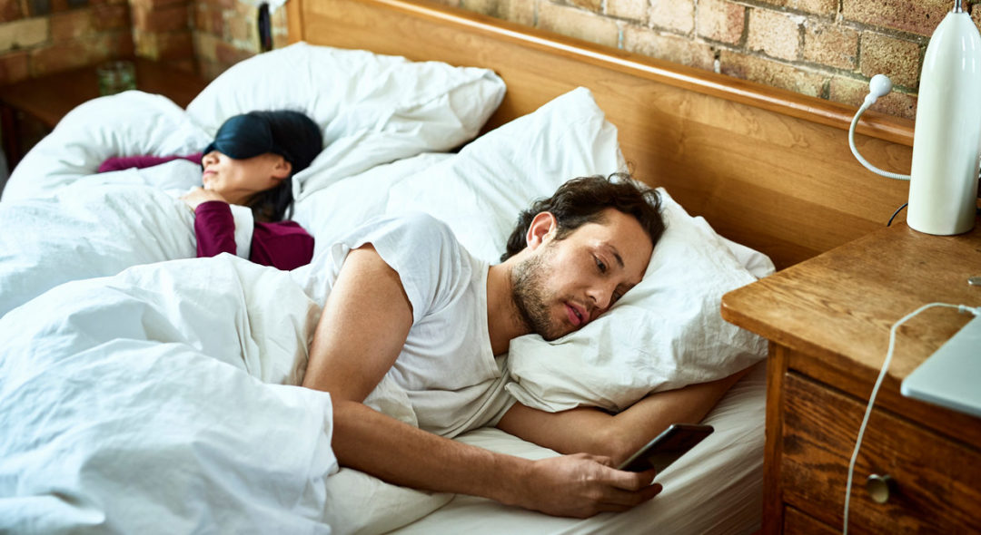 man in bed looking at phone with sleeping woman