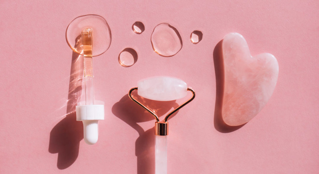 Quartz massager guasha, quartz face roller, pipette and hyaluronan acid drops on pink background
