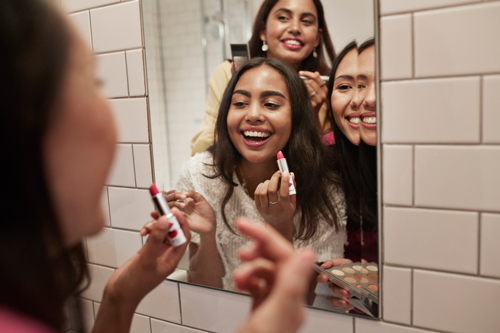 Smiling friends with lipstick looking at mirror