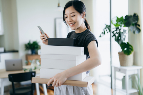 woman with a stack of cardboard boxes delivery package in her arms using smartphone